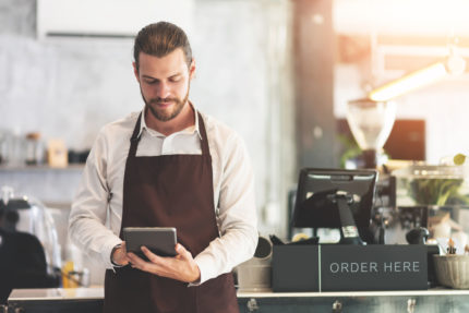 A man in an apron is using a tablet in a coffee shop.