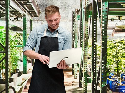 A man in an apron using a laptop in a greenhouse.