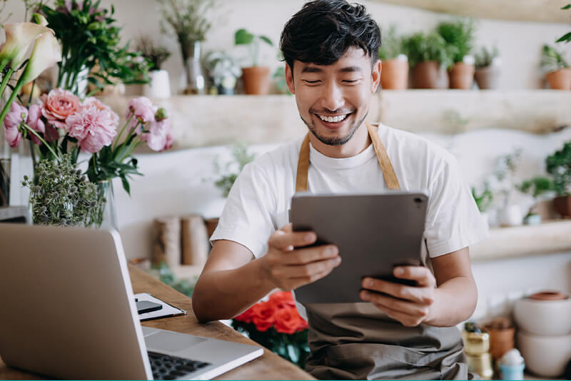 An asian man smiling at a tablet in front of a flower shop.