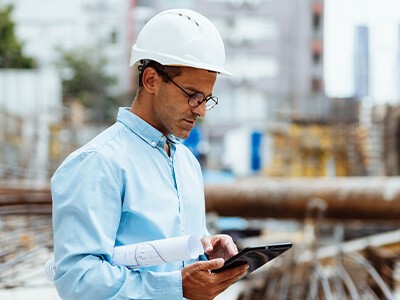 A man wearing a hard hat is holding a tablet while standing on a construction site.