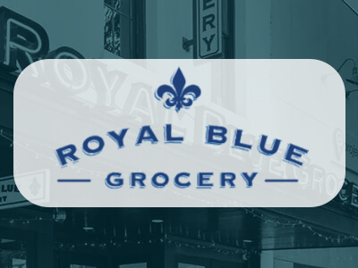 Royal Blue Grocery sign with a fleur-de-lis symbol above the text, set against a blurred background of a grocery storefront.