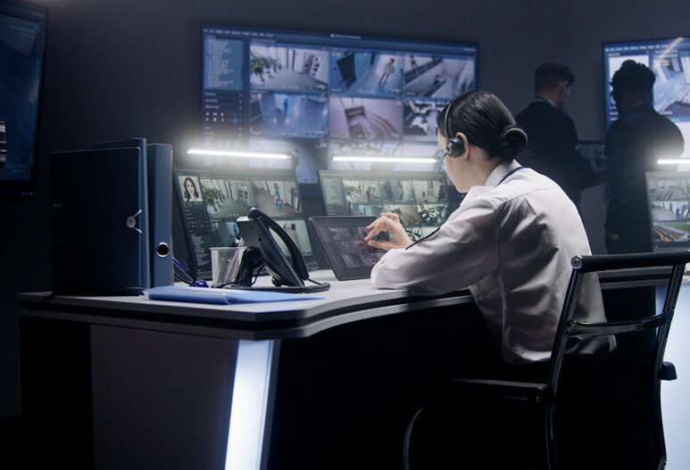 A person in a white shirt monitors multiple security camera feeds on large screens in a control room, with two other people standing in the background.