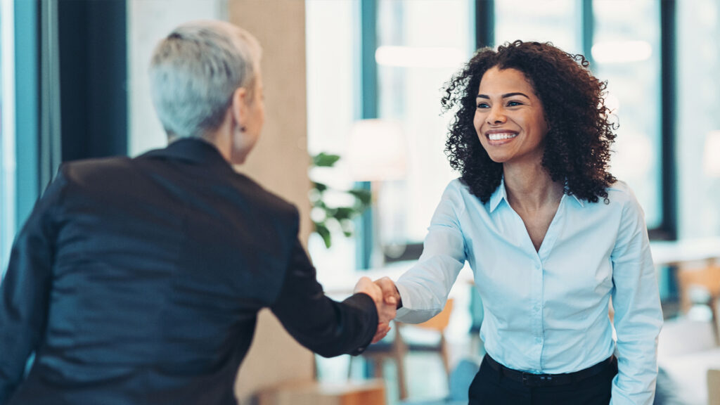 Two professional women shaking hands and smiling in an office setting.