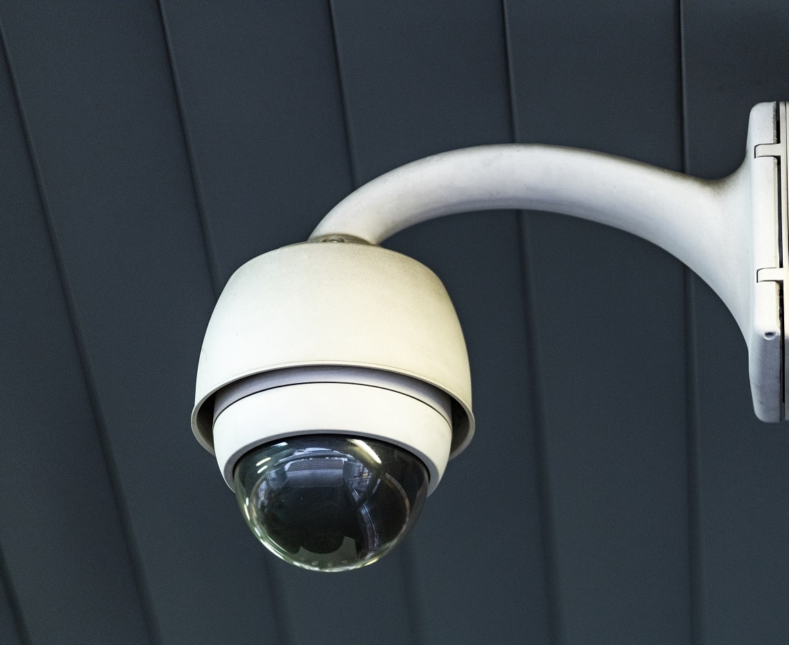 Close-up of a white security camera mounted on a grey wall with horizontal slats.