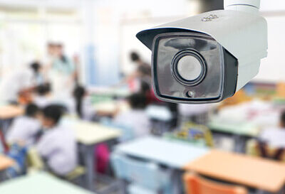 A security camera is mounted on the ceiling in a classroom, monitoring students who are seated at desks.