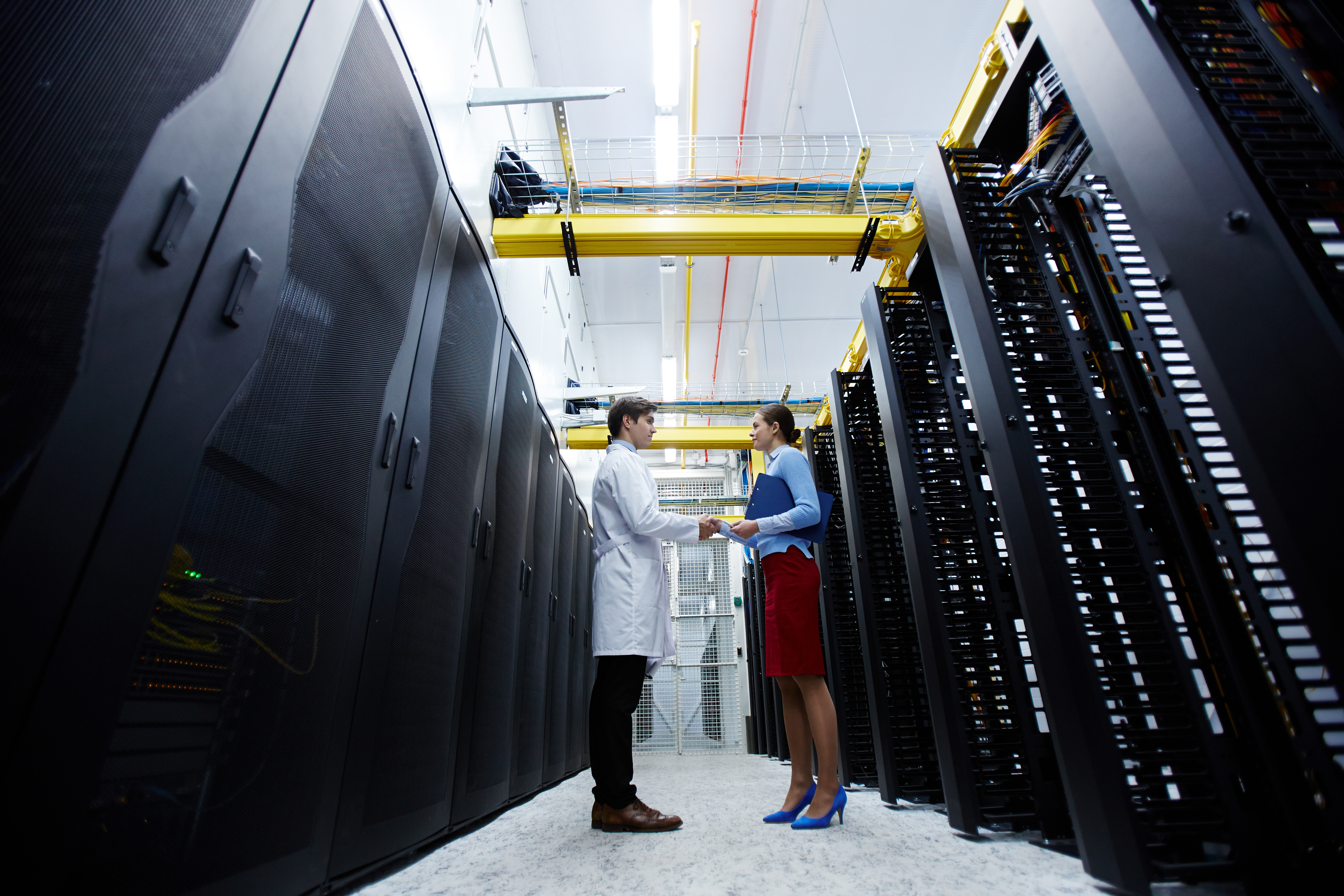 Two young technicians of data center consuting about working points at meeting while standing in aisle of storage room