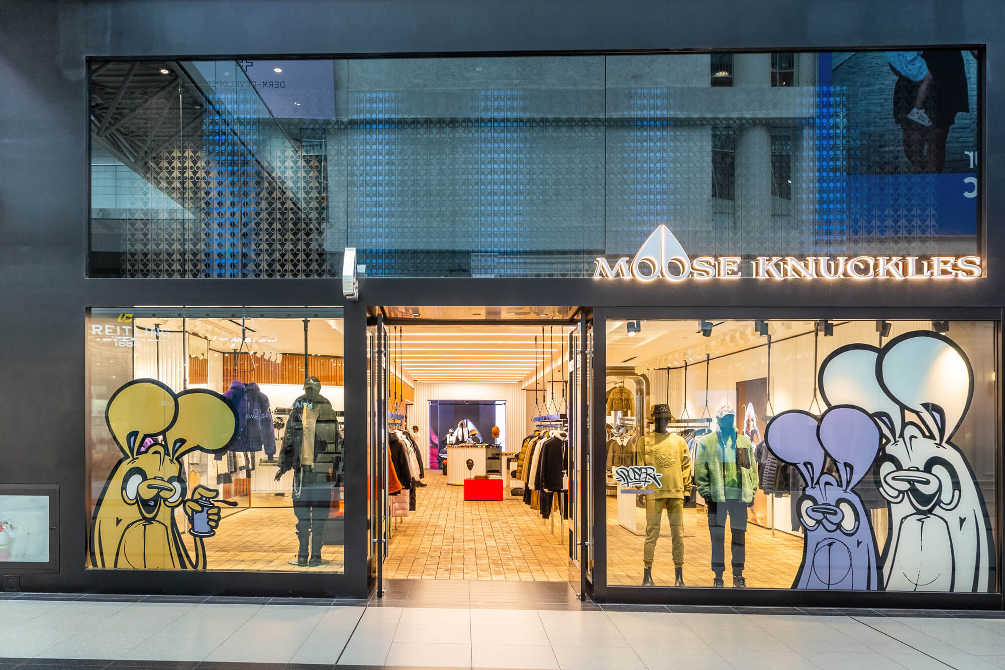 Storefront of Moose Knuckles featuring large windows displaying clothing items. The store’s signage is illuminated, and colorful graffiti art is visible on the lower part of the windows.