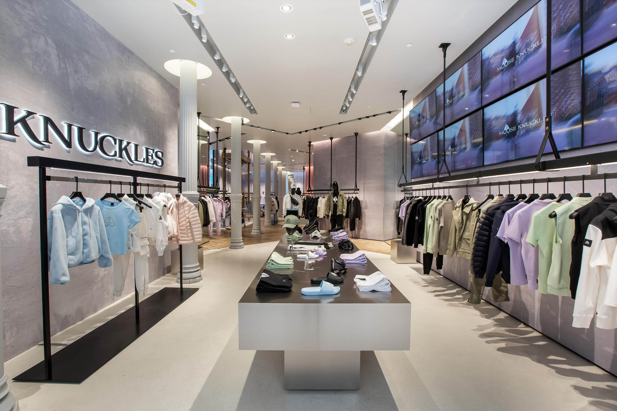 Modern clothing store interior with racks of various garments on both sides and folded clothes on a central display table. Multiple screens line the right wall, displaying imagery.