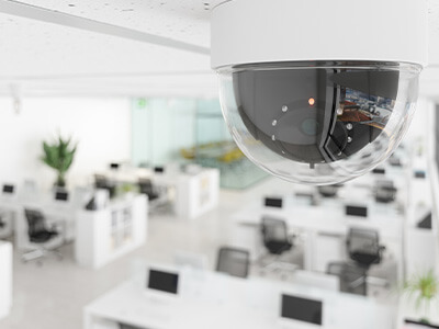 A ceiling-mounted security camera overlooks a modern, open-plan office space with desks, computers, and chairs.