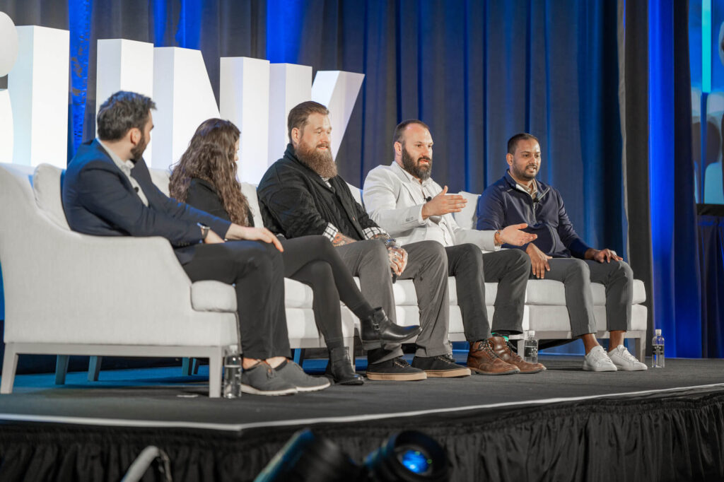 Five individuals are sitting on a stage panel discussing, with one person gesturing as they speak. The backdrop features large white letters.