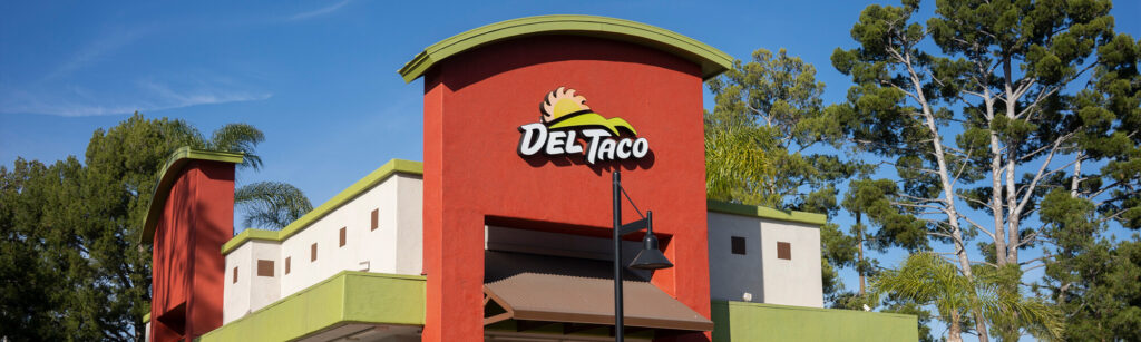 Image of the exterior of a Del Taco restaurant with a red and white building, green accents, and the Del Taco logo prominently displayed on the facade. Trees are visible in the background.