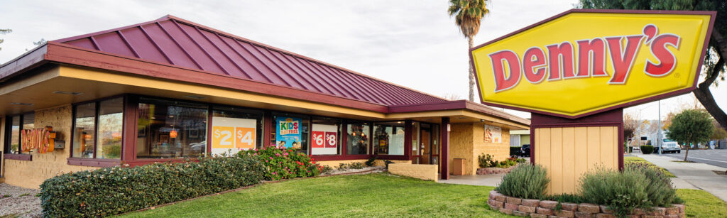 A Denny's restaurant with a yellow and red sign, featuring large windows, a brown-trimmed roof, and neatly landscaped surroundings.