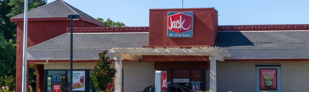 A Jack in the Box fast-food restaurant with a red and gray exterior and visible signage. The entrance has a covered patio area, and promotional posters are displayed in the windows.