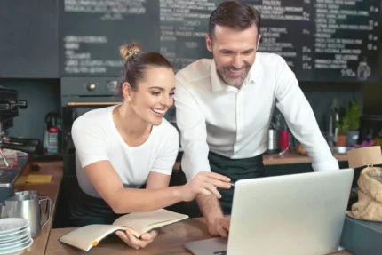 Two people in a cafe look at a laptop screen. One holds a notebook. Chalkboard menu and coffee equipment are in the background.