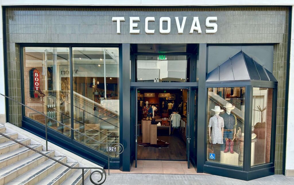 Storefront of Tecovas, featuring western boots and clothing, with mannequins in the display window and a visible interior showing shelves and products.