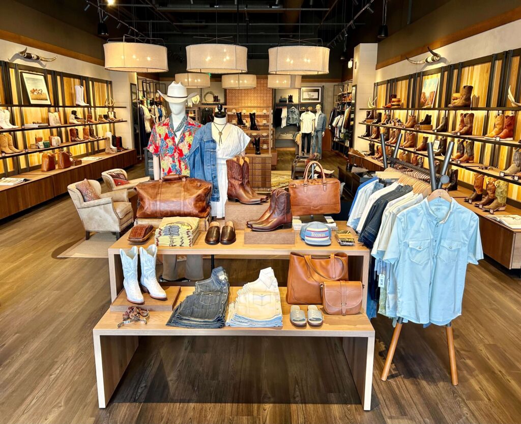 Clothing store interior with wooden flooring. Display tables feature shirts, bags, hats, boots, and jeans. Mannequins showcase casual apparel. Shelves line the walls displaying shoes and accessories.