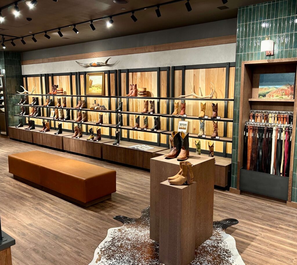A store interior displaying cowboy boots on shelves and stands, with a seating area and a section of belts on the right.