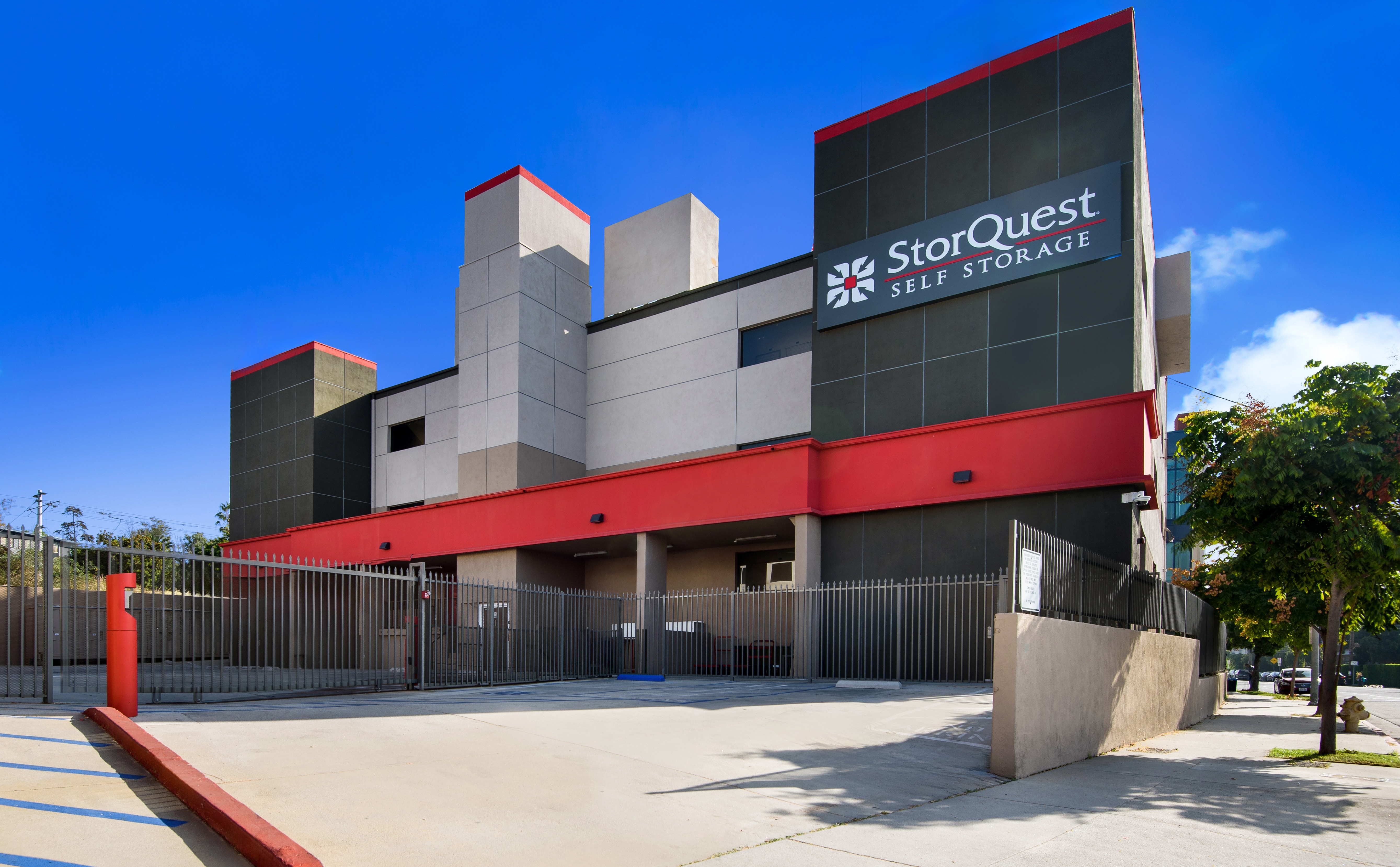 Two-story StorQuest Self Storage building with a red and gray facade, large sign, gated entry, and clear blue sky.