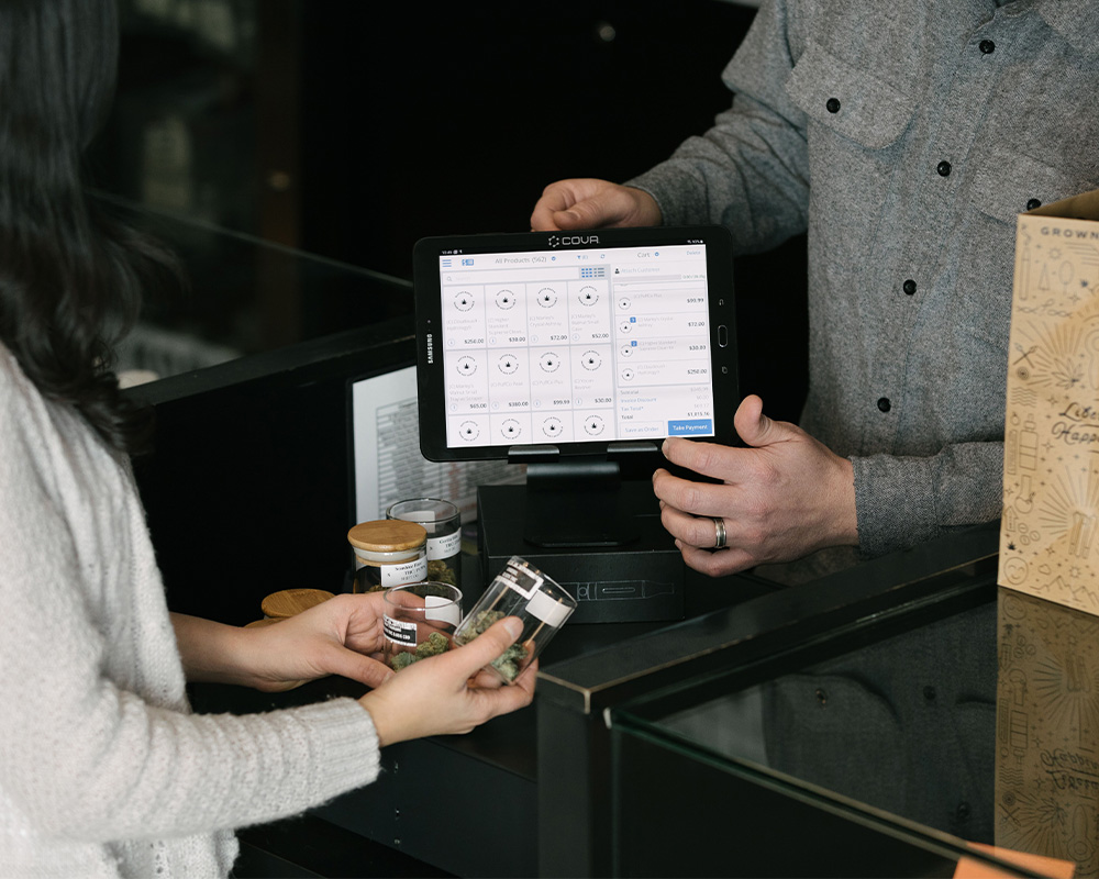 A cashier uses a tablet to assist a customer holding jars, with a box visible in the foreground.