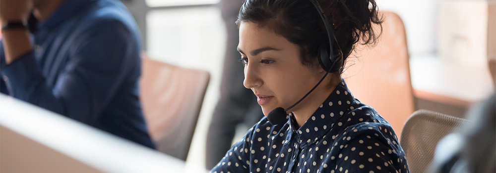 Woman wearing a headset and polka dot shirt works at a computer in an office setting.