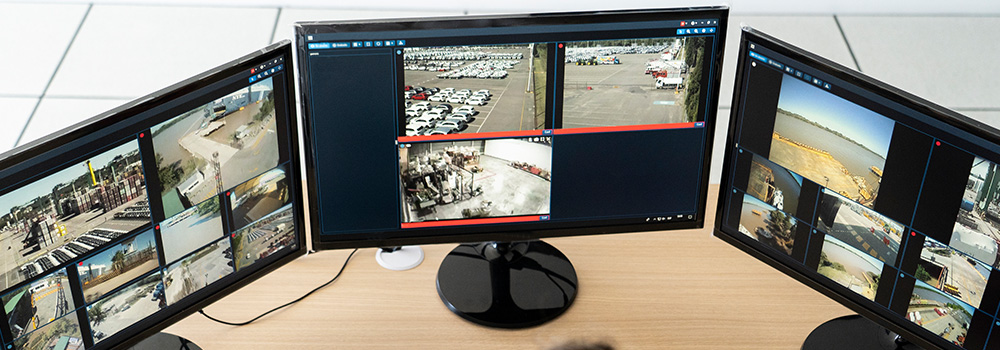 Three monitors on a desk display various surveillance camera feeds showing outdoor and indoor areas.
