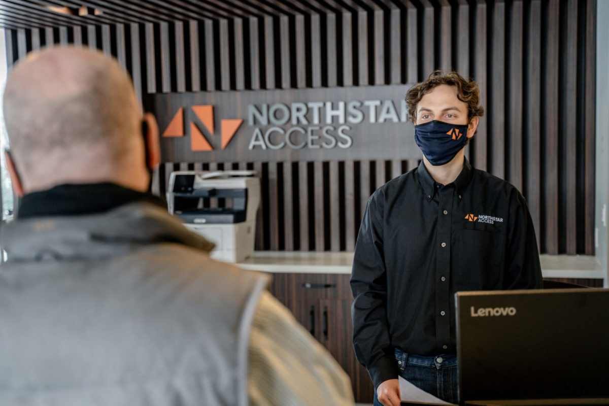 A person in a Northstar Access shirt and mask stands at a reception desk, speaking to another individual with a shaved head. A printer and company logo are visible in the background.