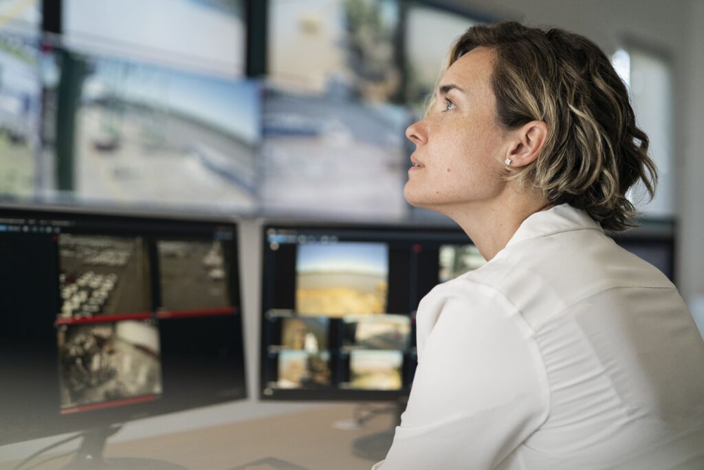 Una persona con una camisa blanca está sentada en un escritorio, mirando varios monitores de vigilancia que muestran imágenes de seguridad.