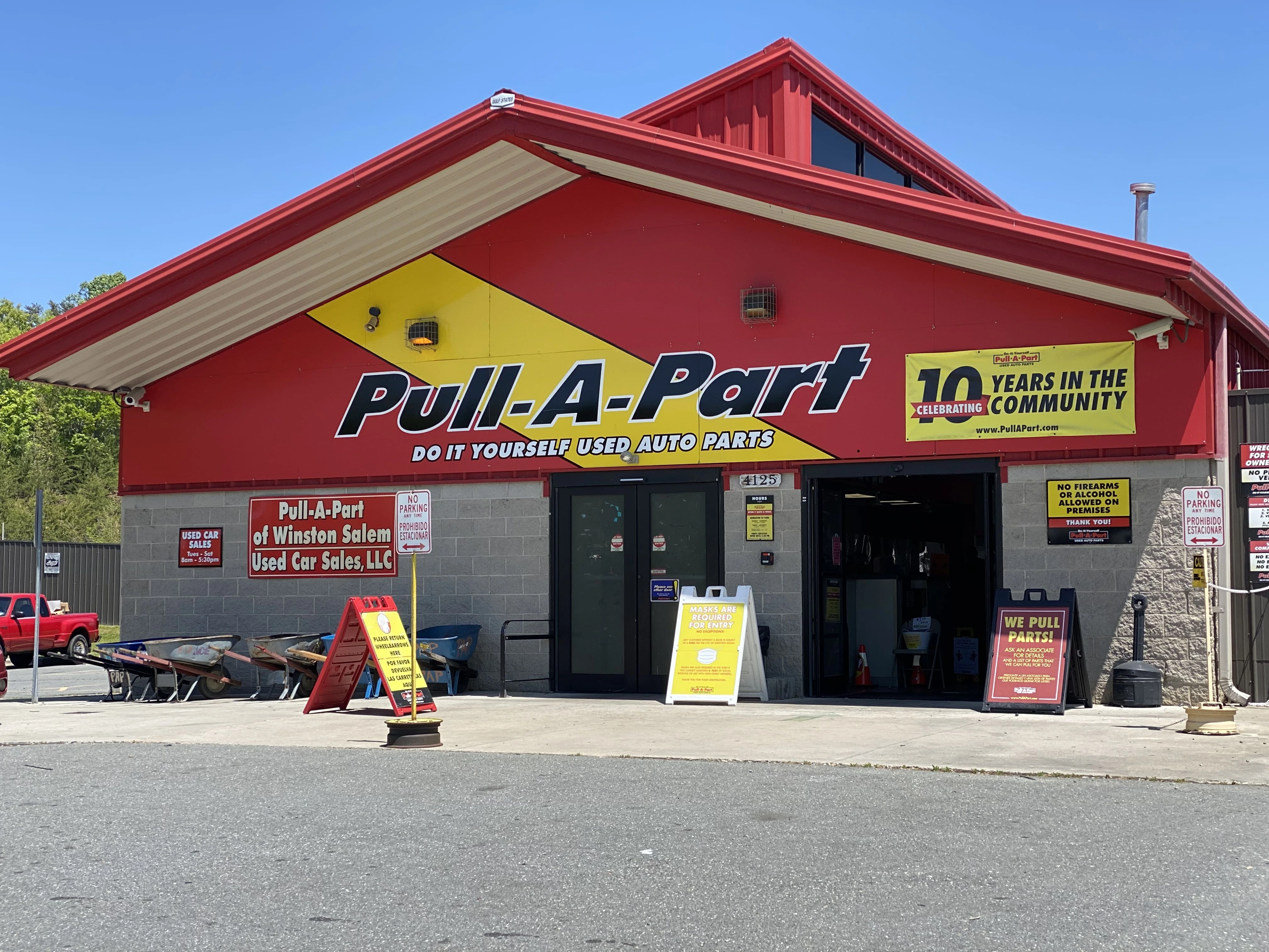 Red Pull-A-Part auto parts store with signs for promotions and do-it-yourself services. Parking lot and wheelbarrows are visible in front.