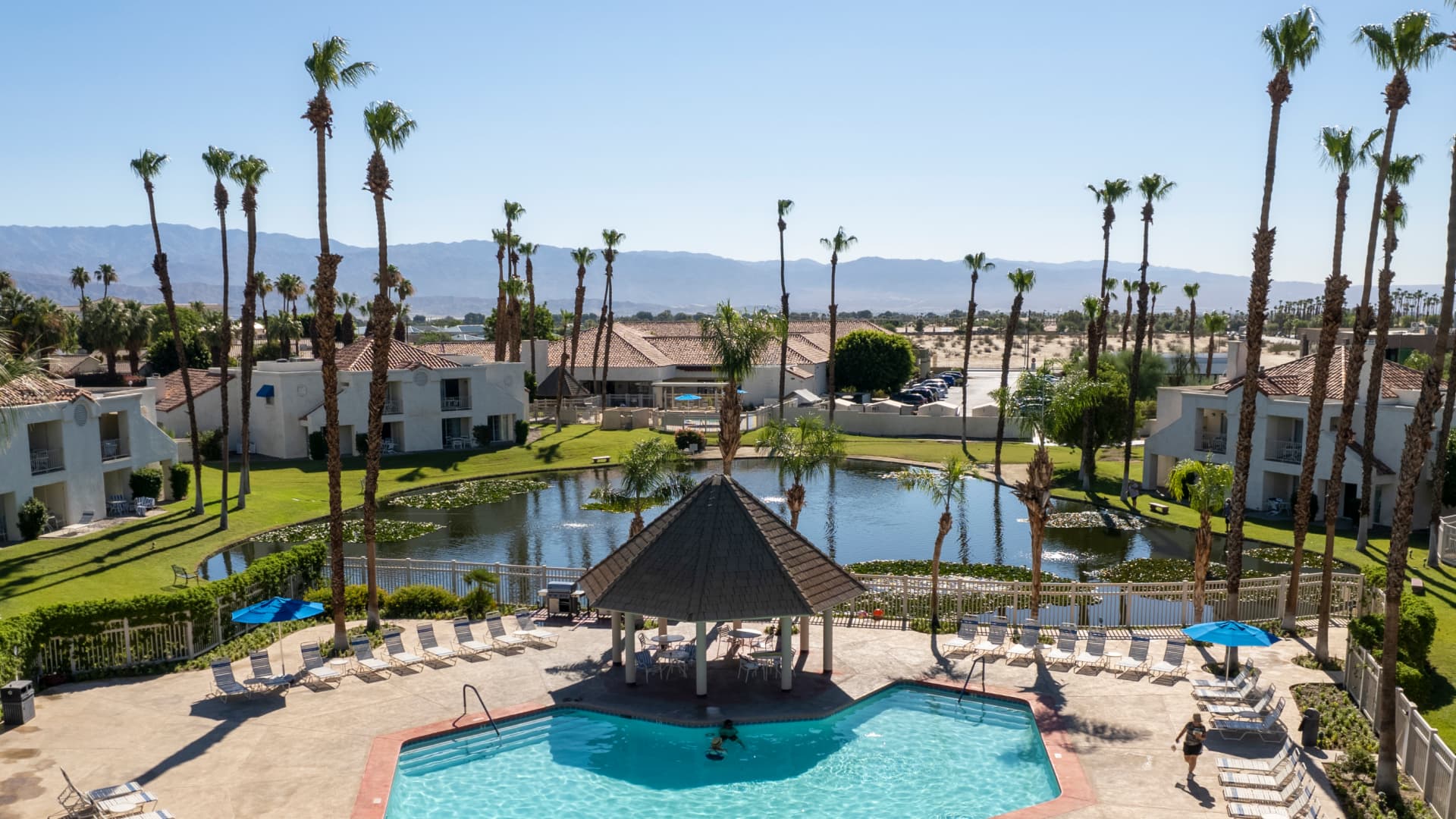 Aerial view of a resort with a central pool shaded by a gazebo, surrounded by sun loungers, palm trees, and a pond. Residential buildings and distant mountains are in the background.