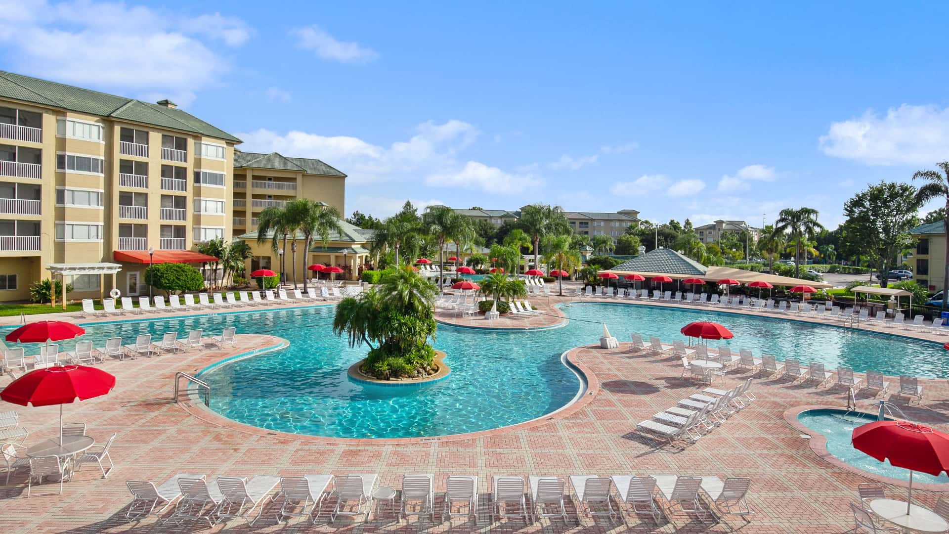 Outdoor pool area at a resort with red umbrellas and numerous lounge chairs, surrounded by palm trees and multi-story buildings.