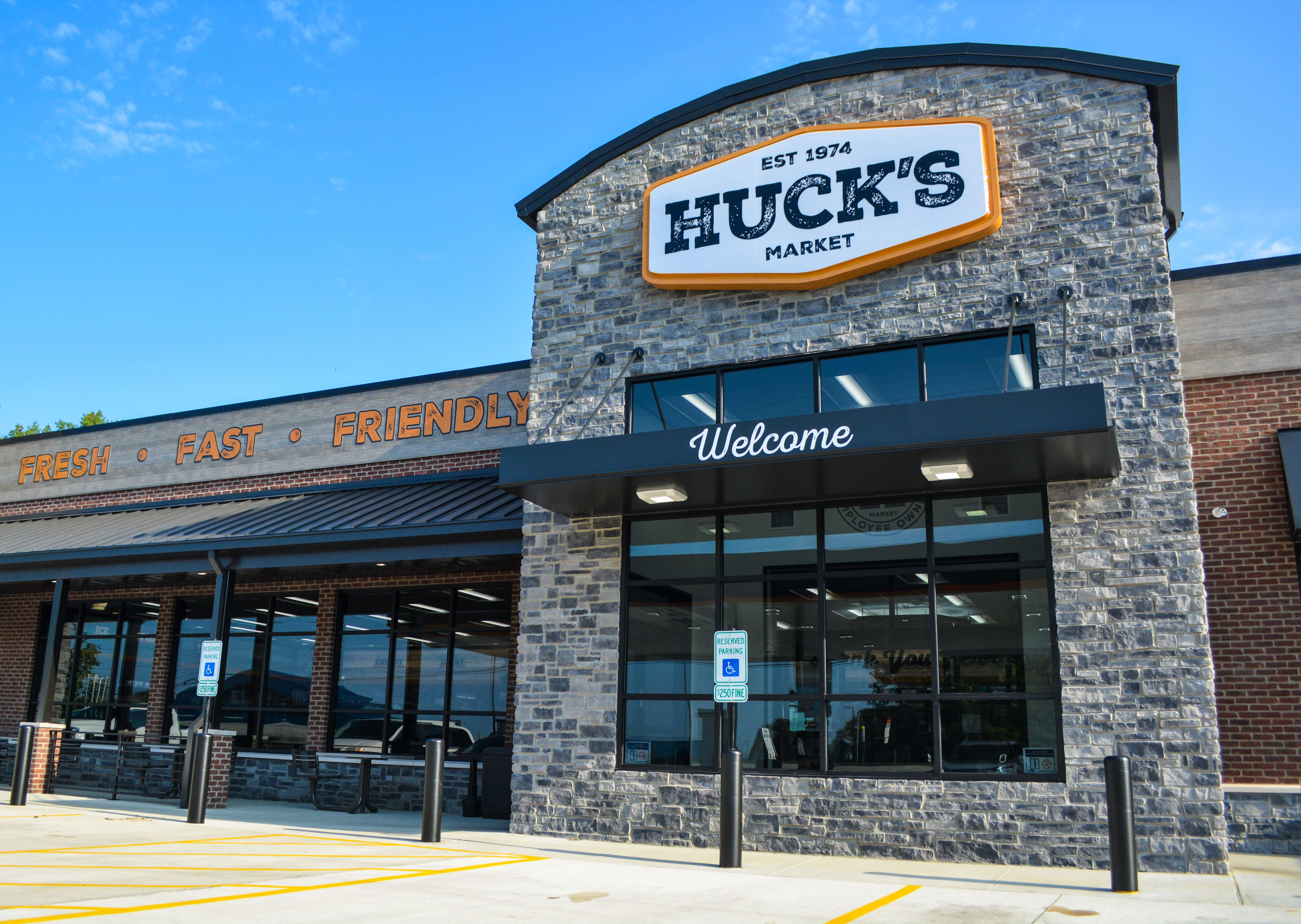 Front view of Huck's Market with a stone facade, large logo sign, and "Welcome" entrance. Signs read "Fresh" and "Fast, Friendly." The building is surrounded by a parking area.
