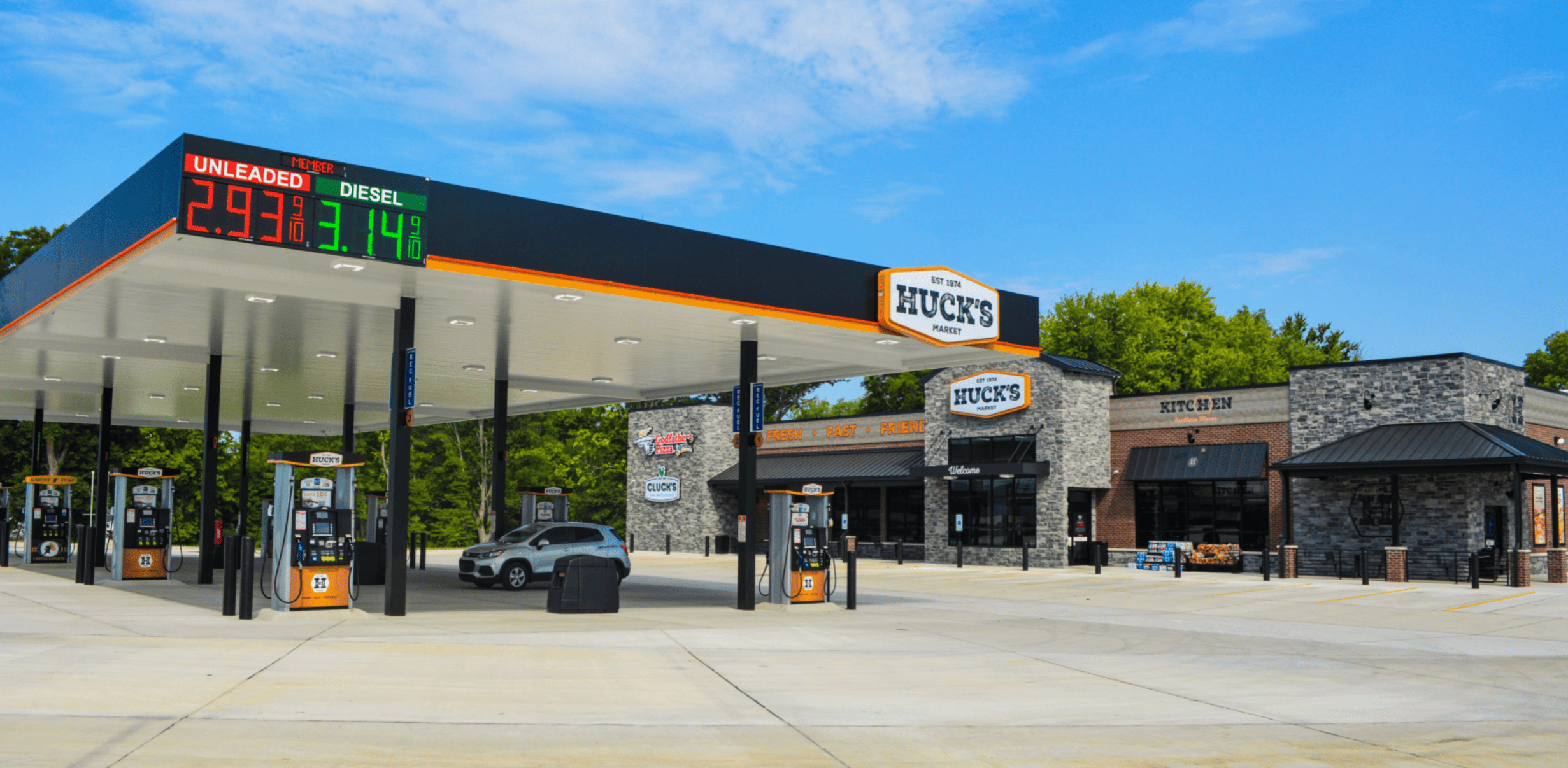 Gas station with multiple fuel pumps under a canopy. Digital sign displays prices for unleaded and diesel. Convenience store in the background, surrounded by trees. Clear blue sky above.