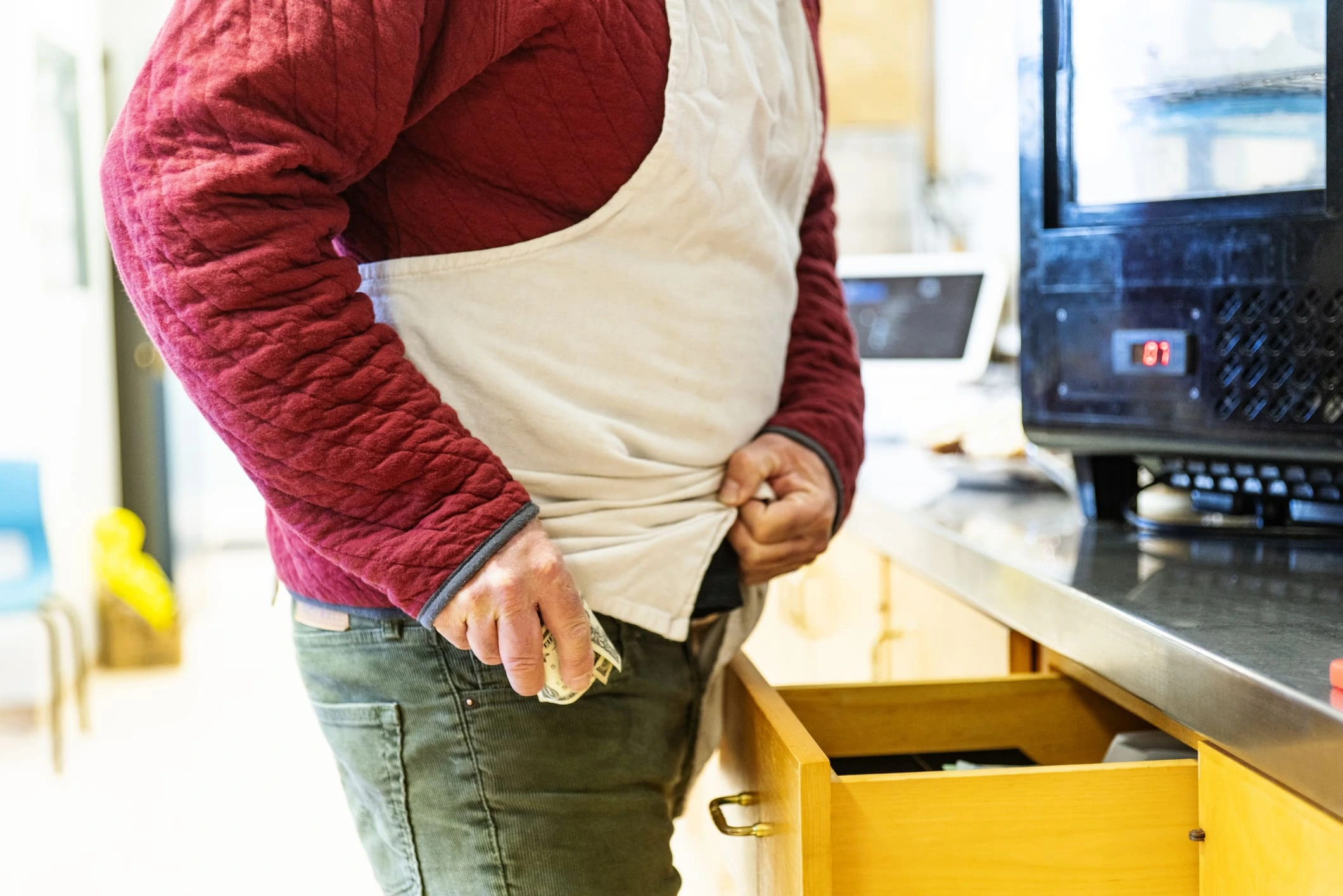 Person in red jacket hides money under shirt while standing near a drawer and an oven in a kitchen.