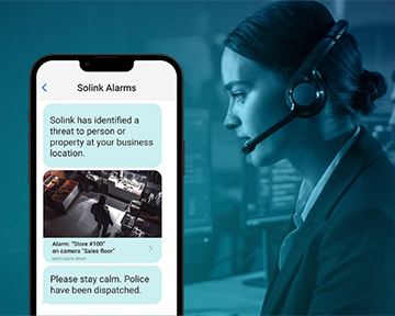 A woman with a headset monitors alerts on her computer while a phone screen displays a security alarm notification and surveillance camera image.