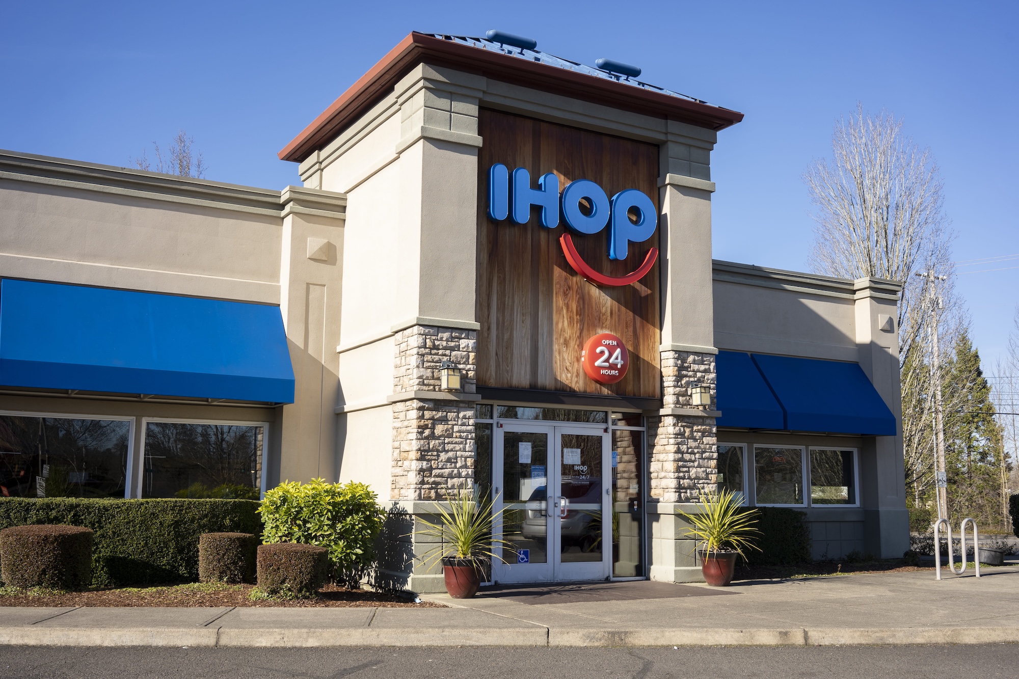 Exterior of an IHOP restaurant with its logo on the building. The entrance has glass doors and the design features stone accents and blue awnings. The area is landscaped with shrubs and plants.
