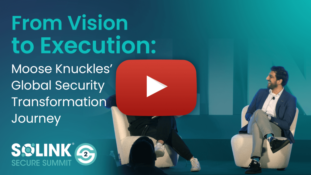 Two men sit on stage at the SOLINK Secure Summit under a projected title, "From Vision to Execution: Moose Knuckles’ Global Security Transformation Journey.