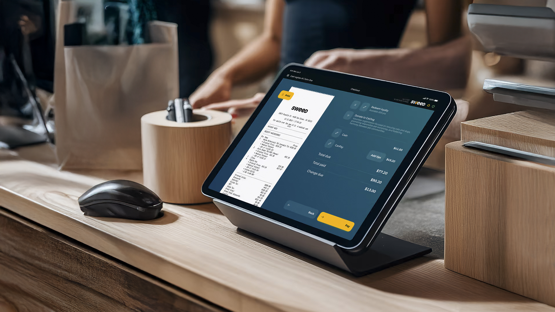 A checkout counter with a digital tablet displaying a receipt, a wireless mouse, a payment terminal, and a paper shopping bag in the background.