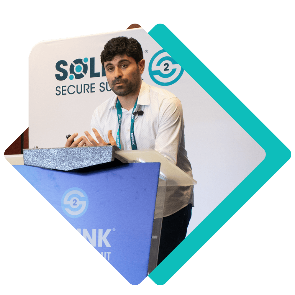 A man stands at a podium giving a presentation at the SOLID Secure Summit, with branding and logos visible in the background.