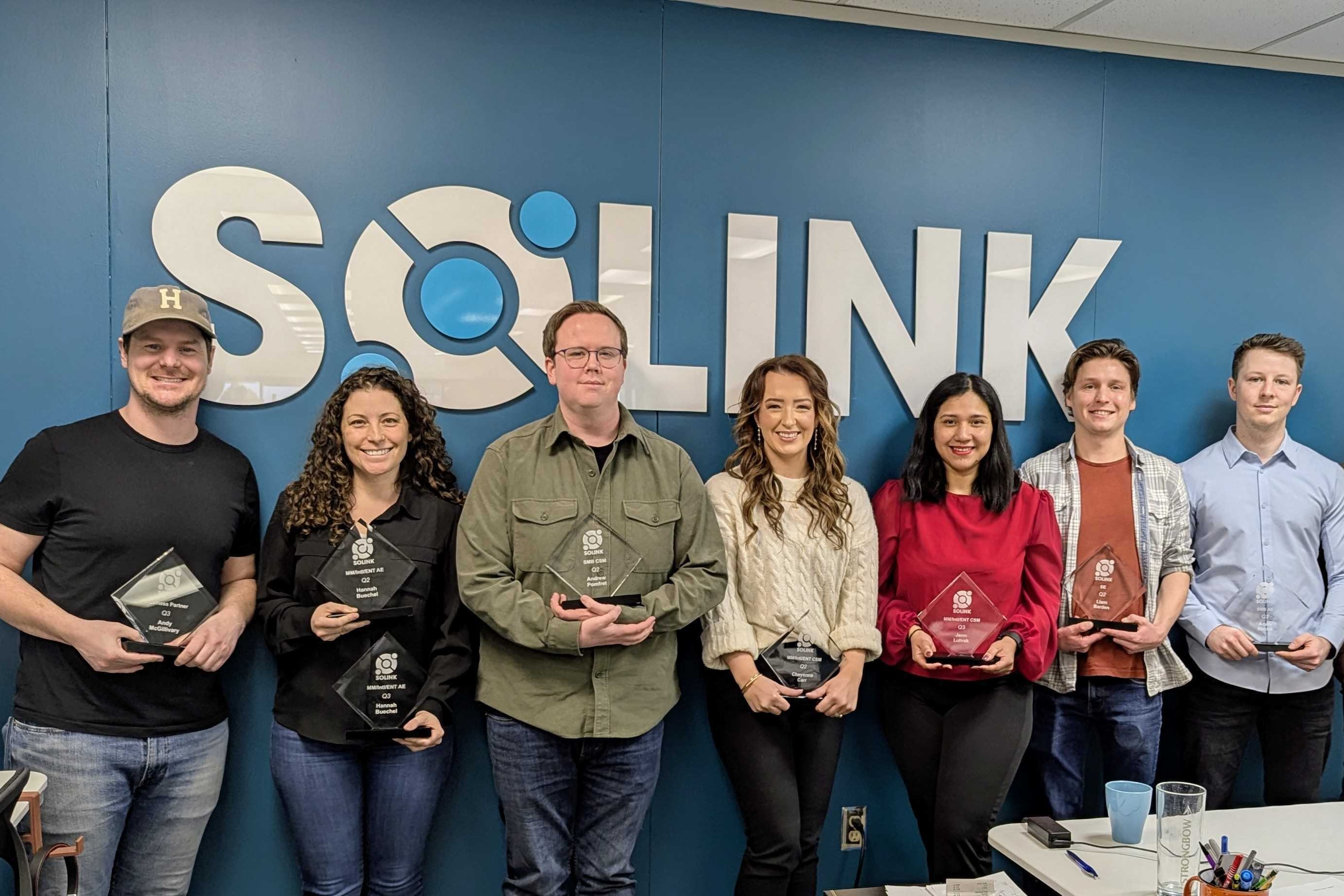 Seven people stand in front of a blue wall with the "SOLINK" logo, each holding an award and smiling at the camera.