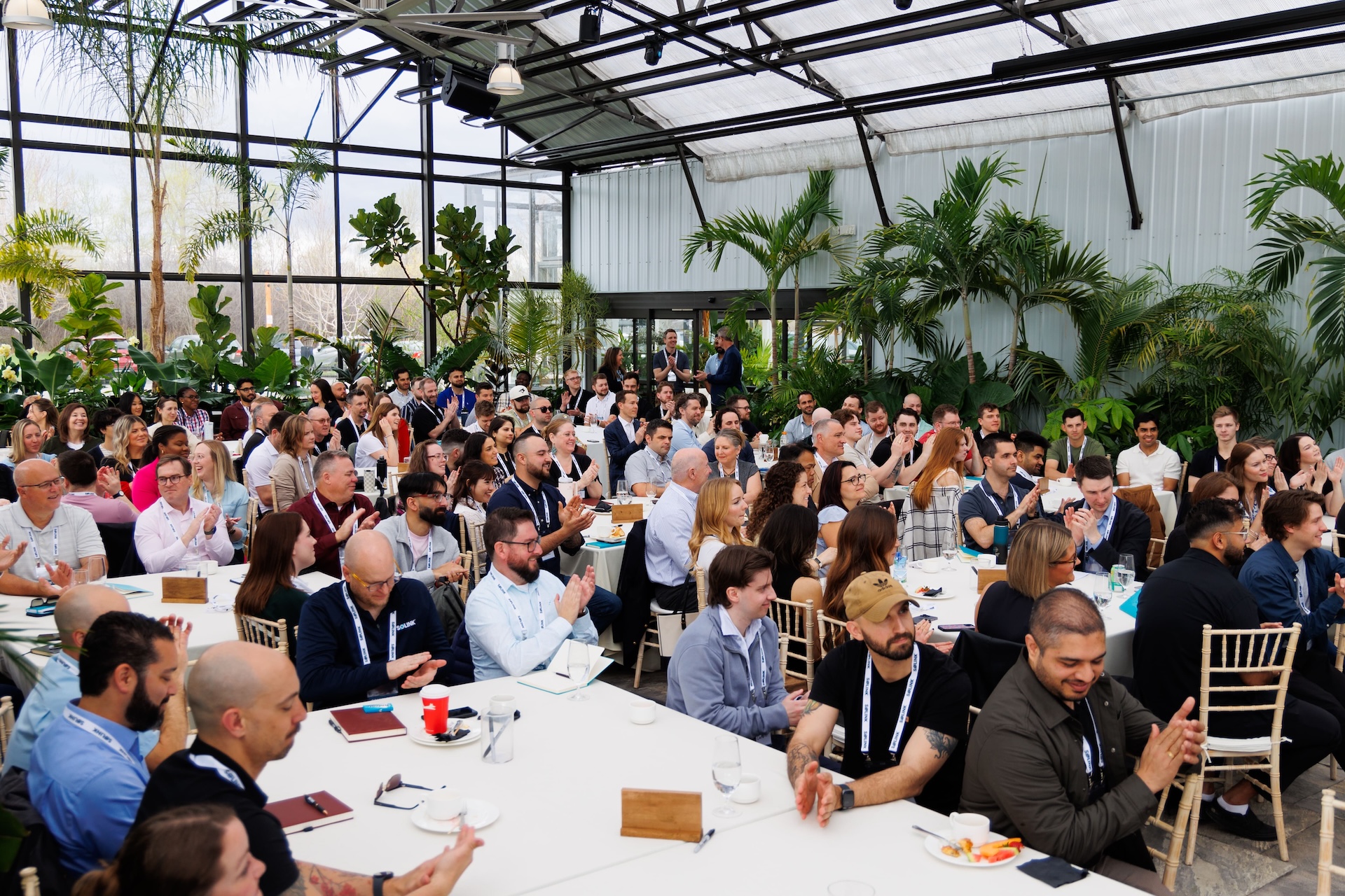 A large group of people seated at round tables in a bright, plant-filled room, listening attentively and applauding during an event or conference.