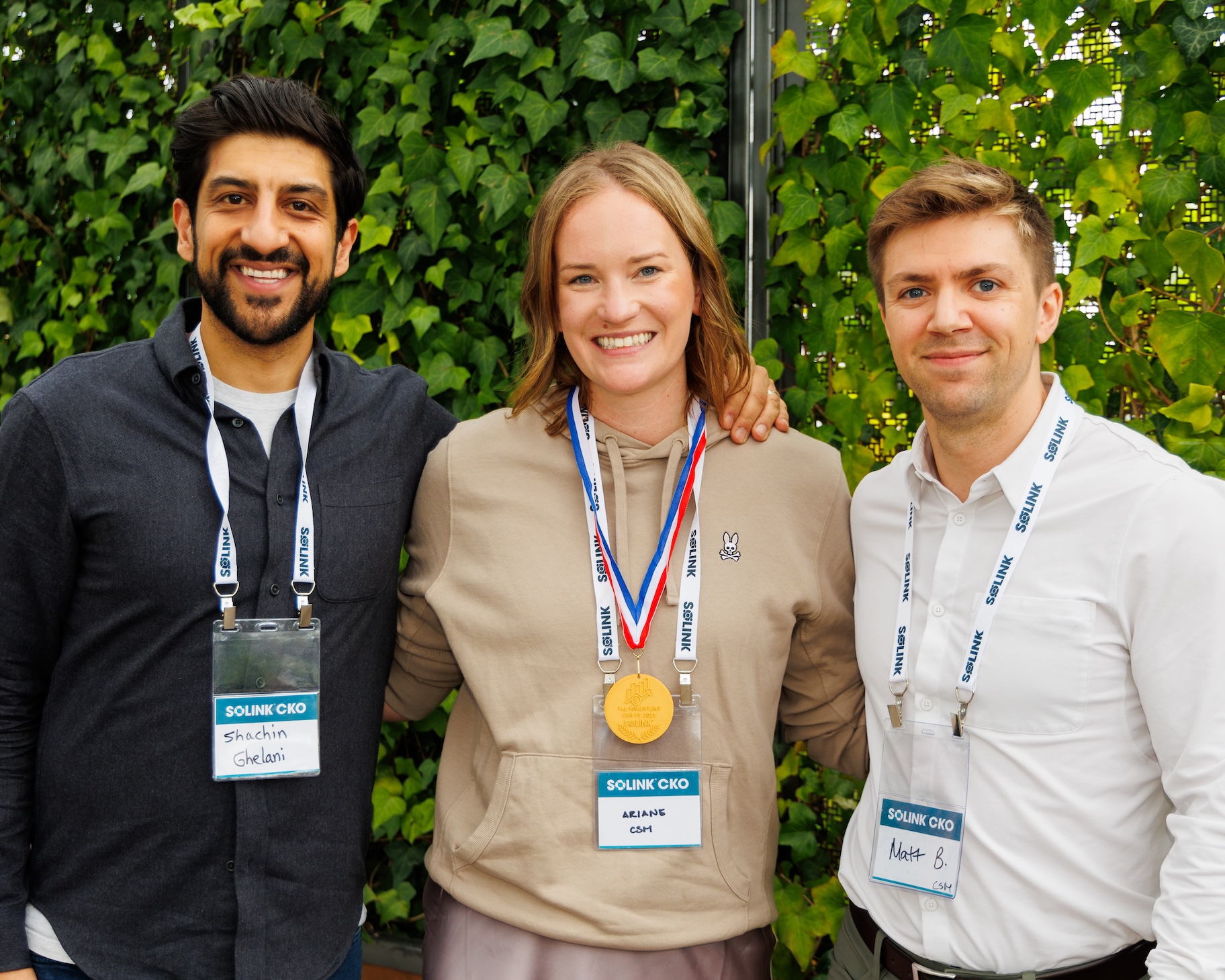 Three people stand together smiling, all wearing conference name badges; the person in the center wears a medal around their neck. Green foliage is visible in the background.