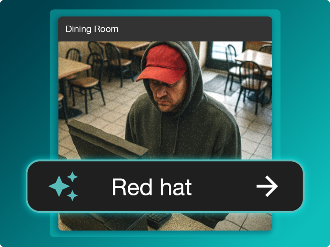 A person in a gray hoodie and red hat sits at a computer in an empty dining room with tables and chairs.