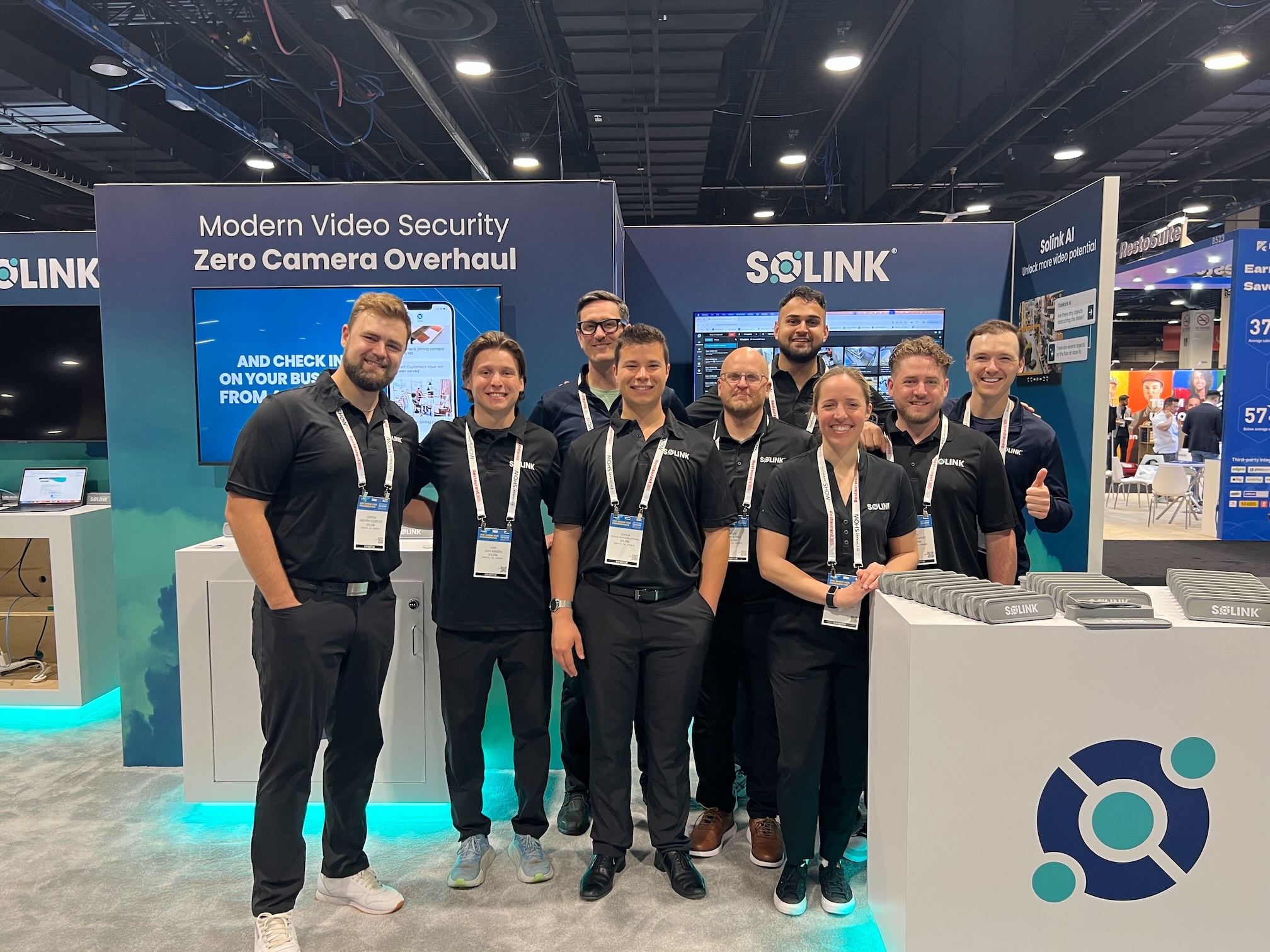 Nine people in matching black shirts stand and smile at a trade show booth featuring Solink branding and video security displays.