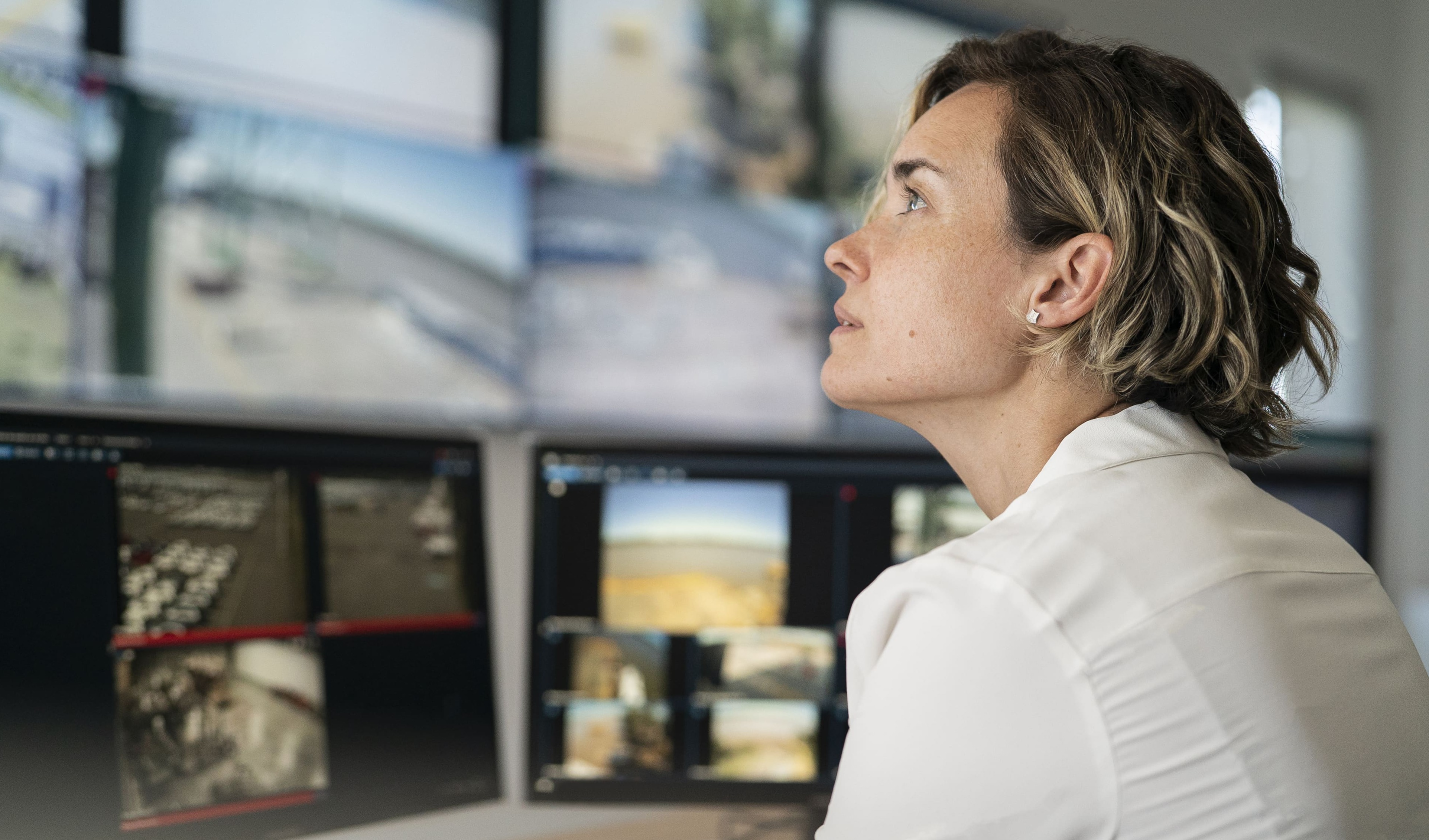 A person in a white shirt closely monitors multiple surveillance screens displaying various camera feeds in a control room.