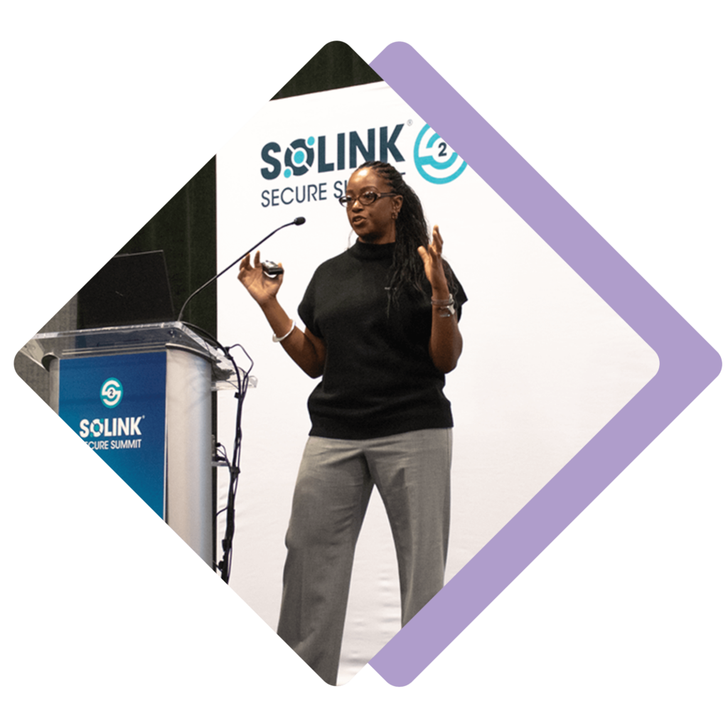 A speaker stands on stage presenting at the Solink Secure Summit, holding a microphone and remote, with a podium and branded backdrop behind her.