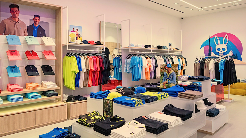 Clothing store interior with folded shirts, pants, and t-shirts displayed on tables and shelves, colorful wall art, and a mannequin wearing casual attire.