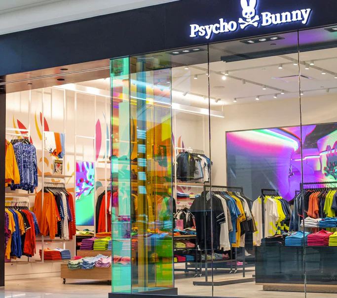 Psycho Bunny clothing store exterior with glass walls, colorful clothing displays, and bright interior lighting inside a shopping mall.