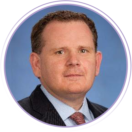 A man with short hair and a light-colored shirt is sitting indoors, looking at the camera. The image has a circular purple border.
