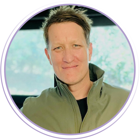 A man with short brown hair and light eyes smiles at the camera, wearing a green jacket. There is a blurred indoor background and a purple circular frame around the image.