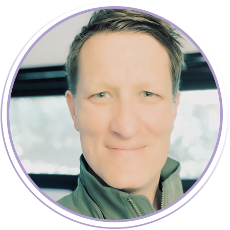 A man with short brown hair and light eyes smiles at the camera, wearing a green jacket. There is a blurred indoor background and a purple circular frame around the image.